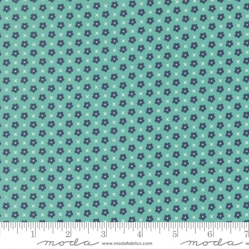 The image is a fabric with a repeating pattern of small blue flowers and white x's. The pattern is arranged in a diagonal direction, creating a sense of movement across the fabric. The fabric appears to be made of a material with a light blue-green hue, and there is a ruler visible at the bottom of the image, indicating the fabric's size.